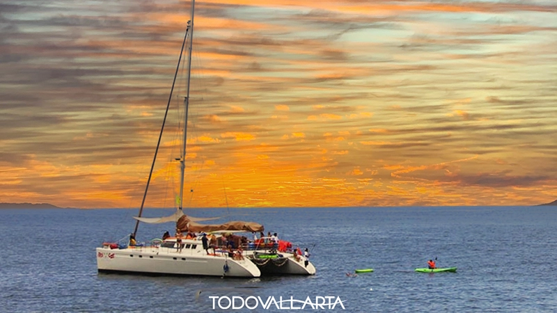 The 56-foot Ally Cat glides at sunset with two kayakers nearby during a catamaran charter 56.